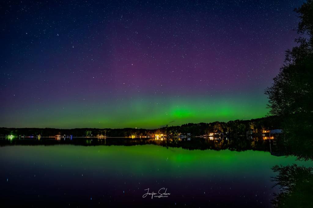 Photo of purple and pulsing green Aurora over a lakeshore in Northern Michigan with the houses on the lake lighting up.